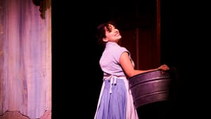 Campos in an apron, long blue skirt, patterned top, holds a farm bucket, smiling, turning, looking behind over her shoulder