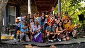 About 2 dozen diverse smiling people, many holding jaranas, pose together on a large porch in summertime.