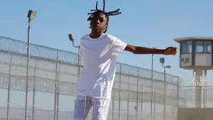 A Black boy in white T and shorts, his arms extended out, in movement in an outdoor incarceration facility area