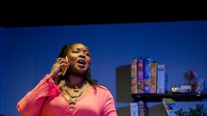 Scene from ‘SOON.’ Actor Ebony Pullum, a Black woman, in a pink shirt & striped pants, talks on the phone in a small kitchen