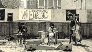 A band of 3, drums, cello, and keys, speakers on tripods on an outdoor stage, a poster with WEIRDO hand-written on it