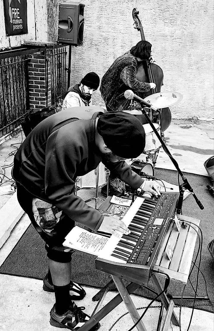 Black & white outdoor photo of the black-hatted musicians playing, bending over keyboard, drums, and upright bass.