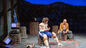 On a set of tree stumps and TV sets, Lally, wearing hockey pads, talks avidly to Gordon, who wears a mustard-colored jacket.