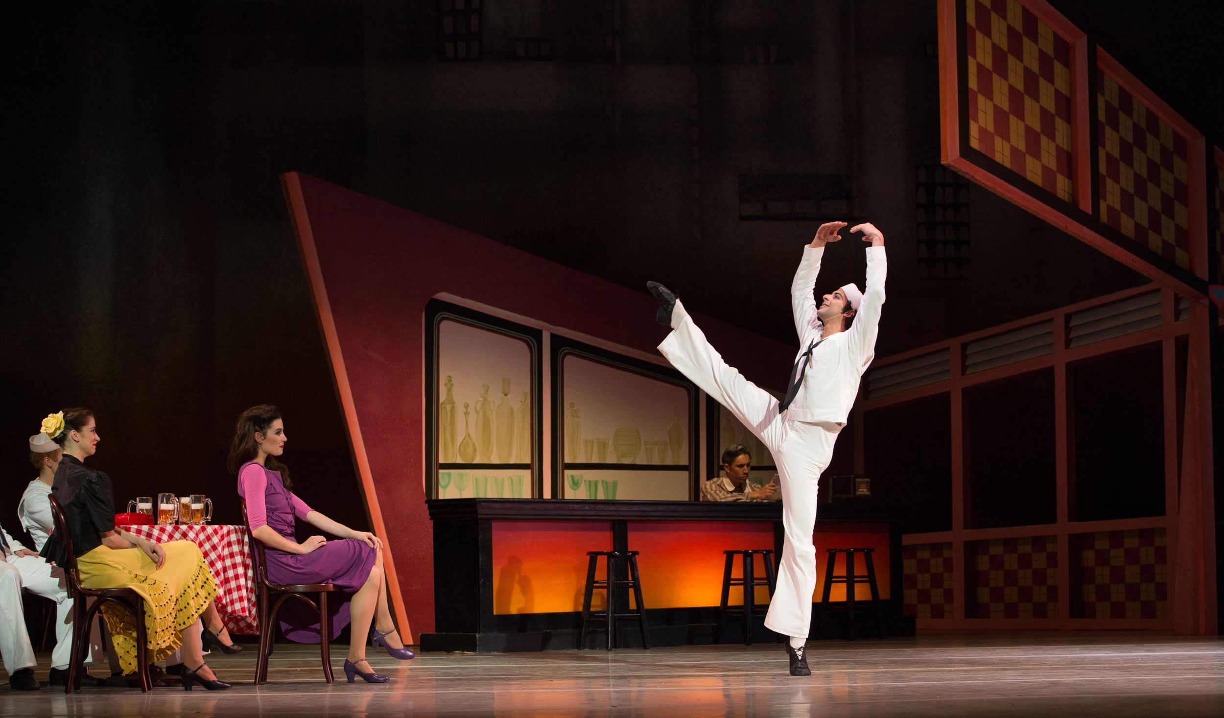 In a midcentury bar setting, Soca, in white sailor suit, poses kicking one leg straight up.