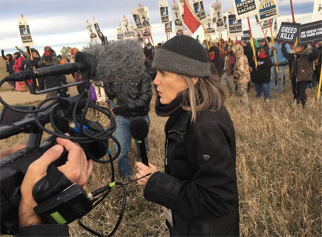 A woman holds a mic, camera facing her, outdoors. A crowd holds protest signs, many say ‘Defend the Sacred.’