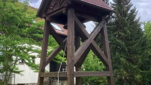 A bell tower with thick brown wooden posts and cross-pieces and a small tiled roof, against summer greenery and a cloudy sky.