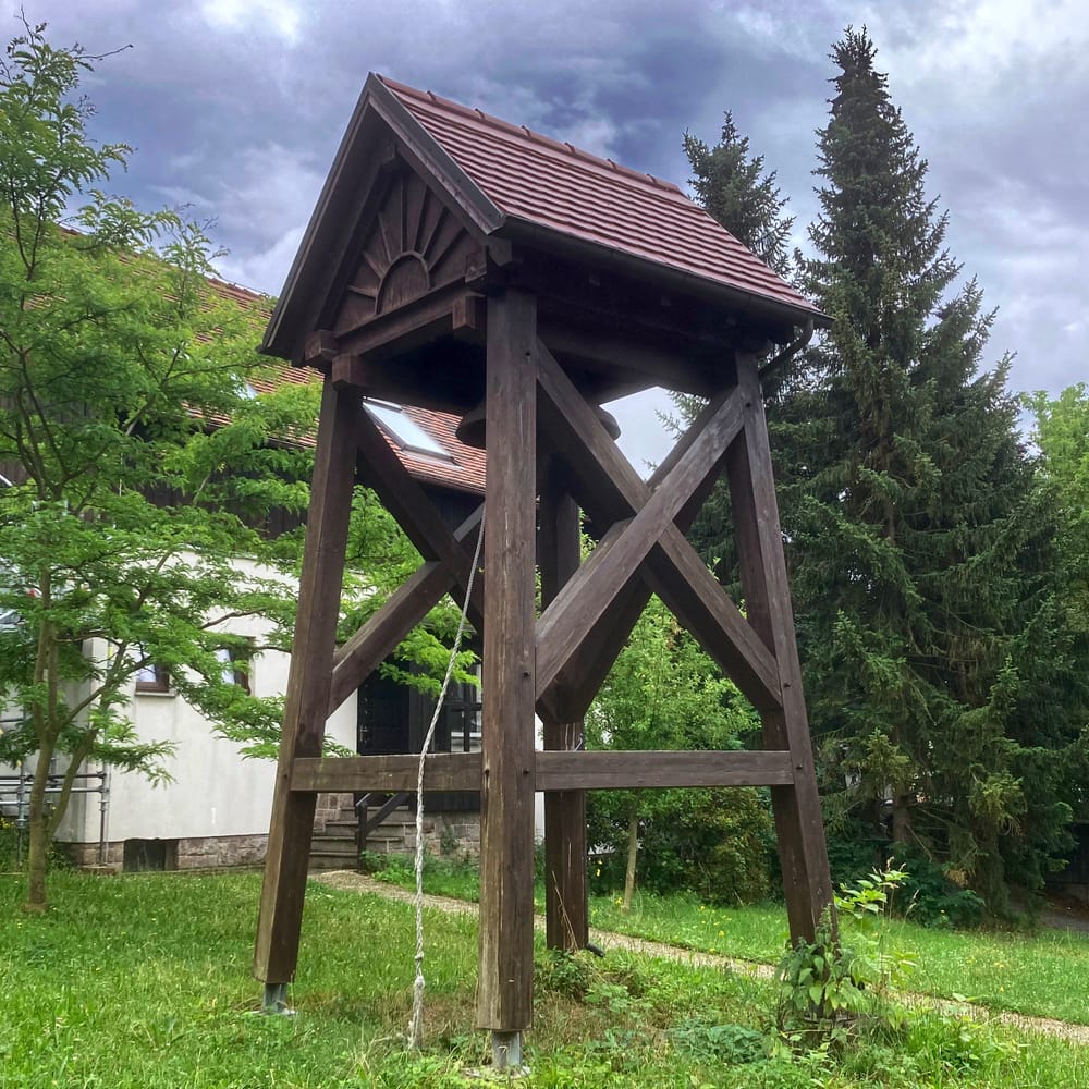 A bell tower with thick brown wooden posts and cross-pieces and a small tiled roof, against summer greenery and a cloudy sky.