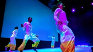 Four dancers move on a blue-lit stage wearing loose white garments with yellow and orange color from the knees to the ankles