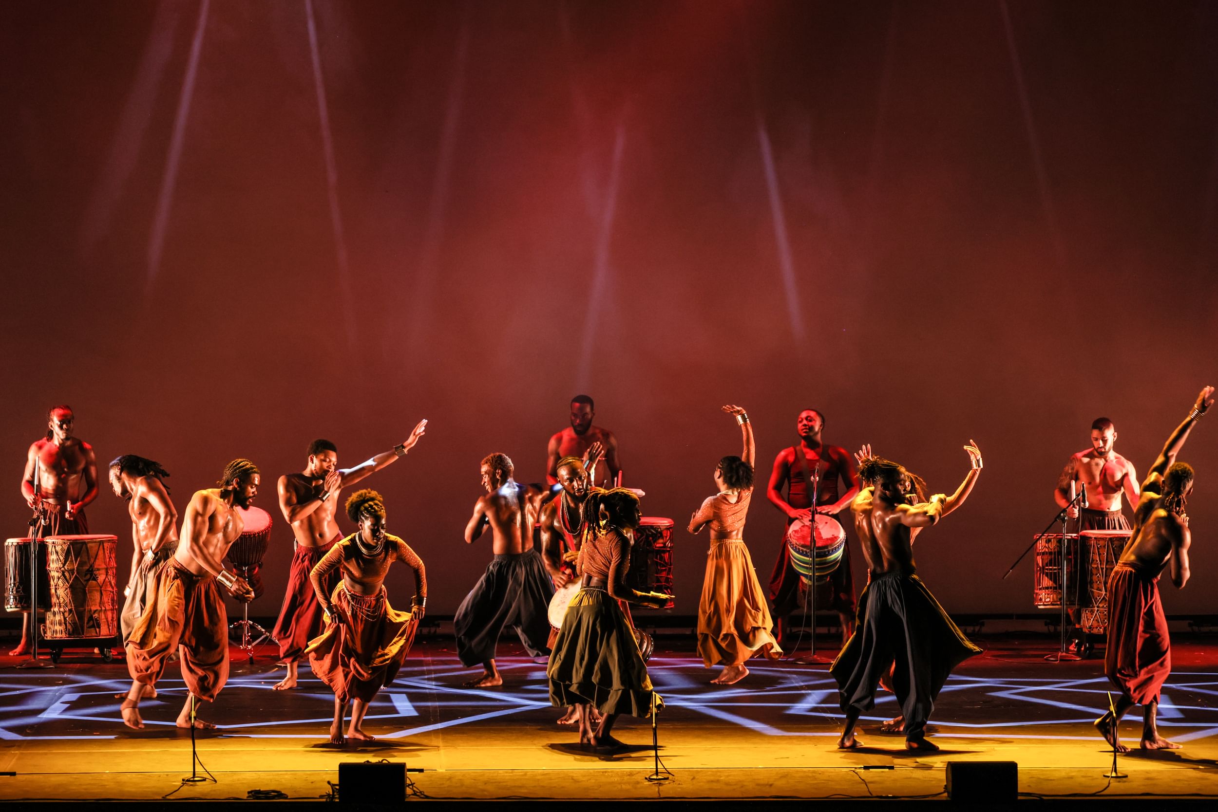 A large group of dancers on stage, with drums in the back and dancers performing on stage; in dresses and harem pants