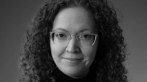 A black and white portrait of Feldman. She wears a sweater over a black sweater with a necklace, glasses, and curly hair
