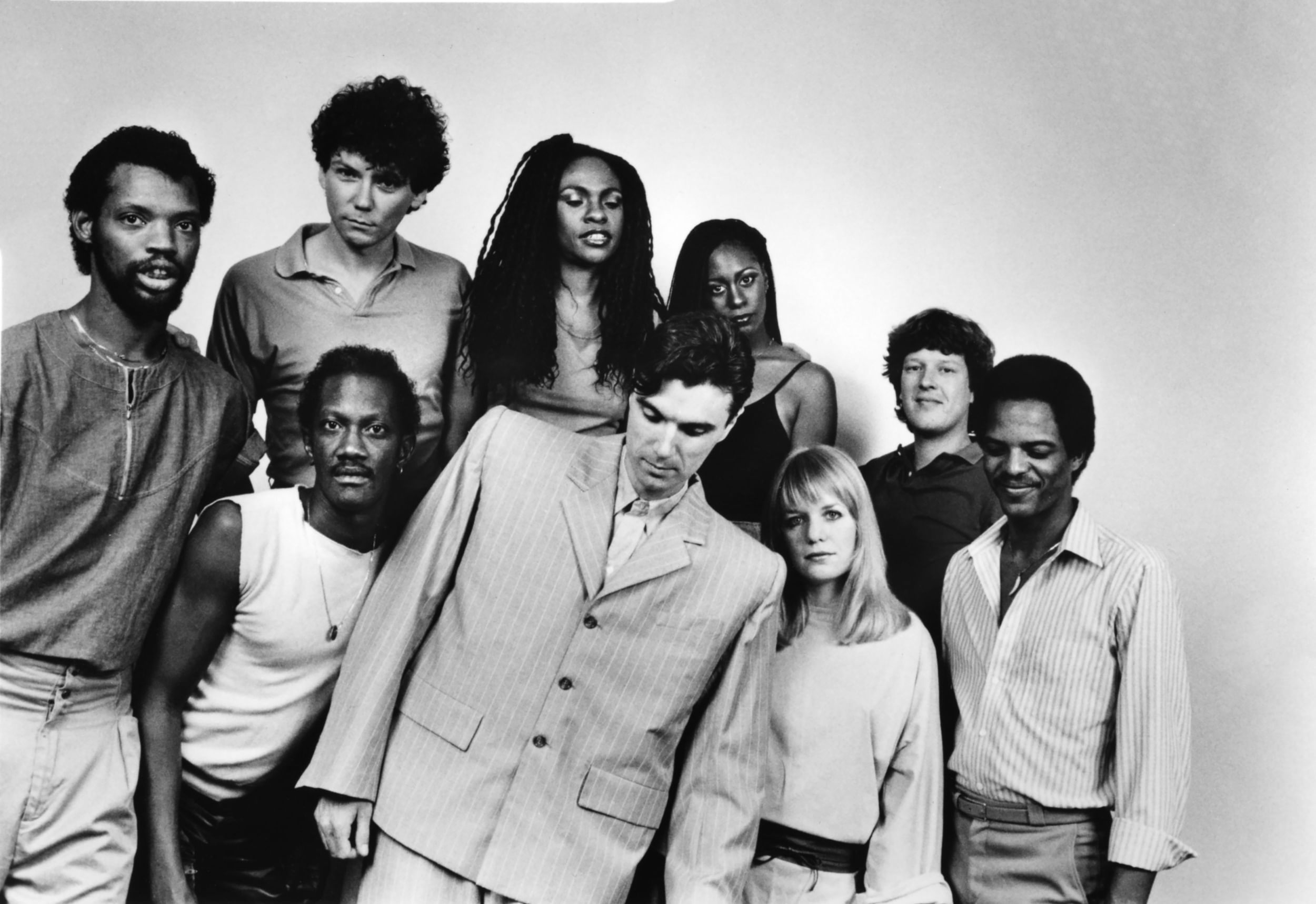 Black & white photo of 9 people posing with different expressions in 1980s garb, David Byrne at center in an oversized suit