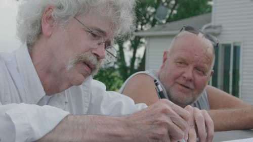 Two white men with white hair sit talking at an outdoor table. One grasps a pen to write something that is out of view.