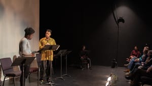 Two Black performers reading from music stands face each other in front of an audience crowding the frame at right.