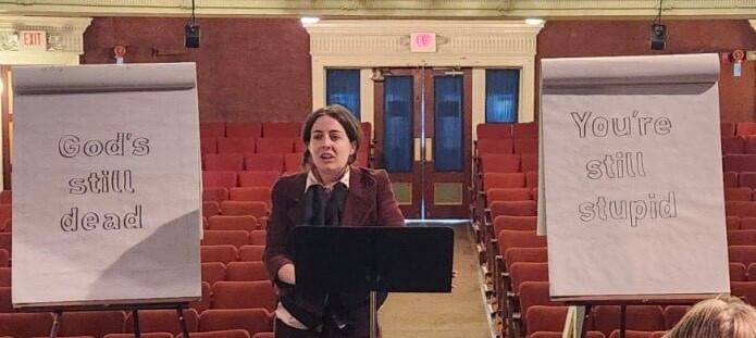 Adams, a white woman in maroon blazer, lectures onstage between signs saying “god’s still dead” and “you’re still stupid"