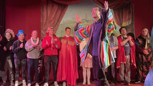 One joyful actor in a rainbow cloak walks in front of about ten other performers in colorful costumes & different expressions
