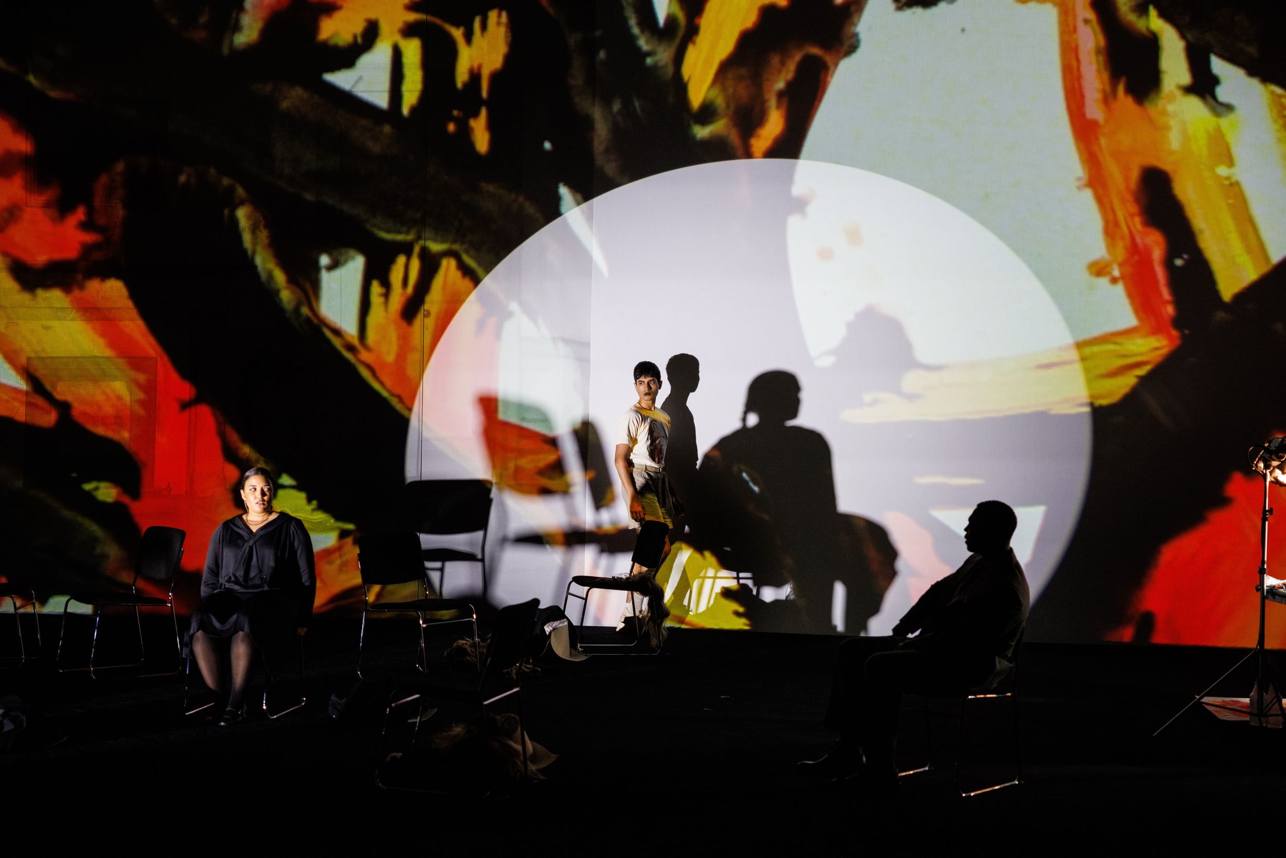 3 actors, 1 in shadow & 1 standing spotlit, arrayed in front of a giant projection of a red, black & yellow abstract painting