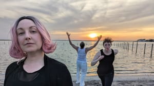 3 cast members, including Ross Beschler with his back to the camera, strike different poses on a bay shore at sunset.
