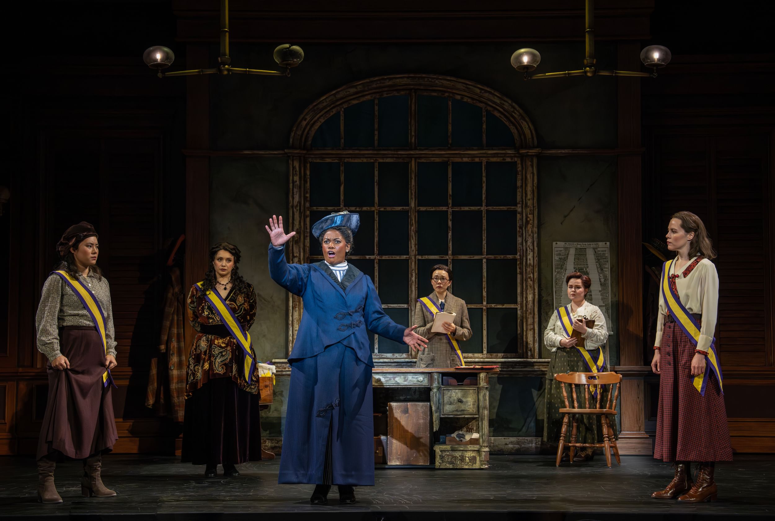 Fulton, a Black woman in a satiny blue dress and hat, sings passionately while five ensemble members look on.