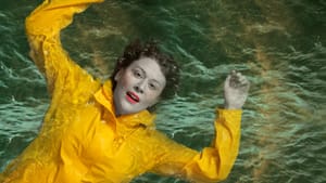 Show logo: Krista Apple, a white actor with short hair, floats in a green sea, wearing a yellow slicker and red lipstick.