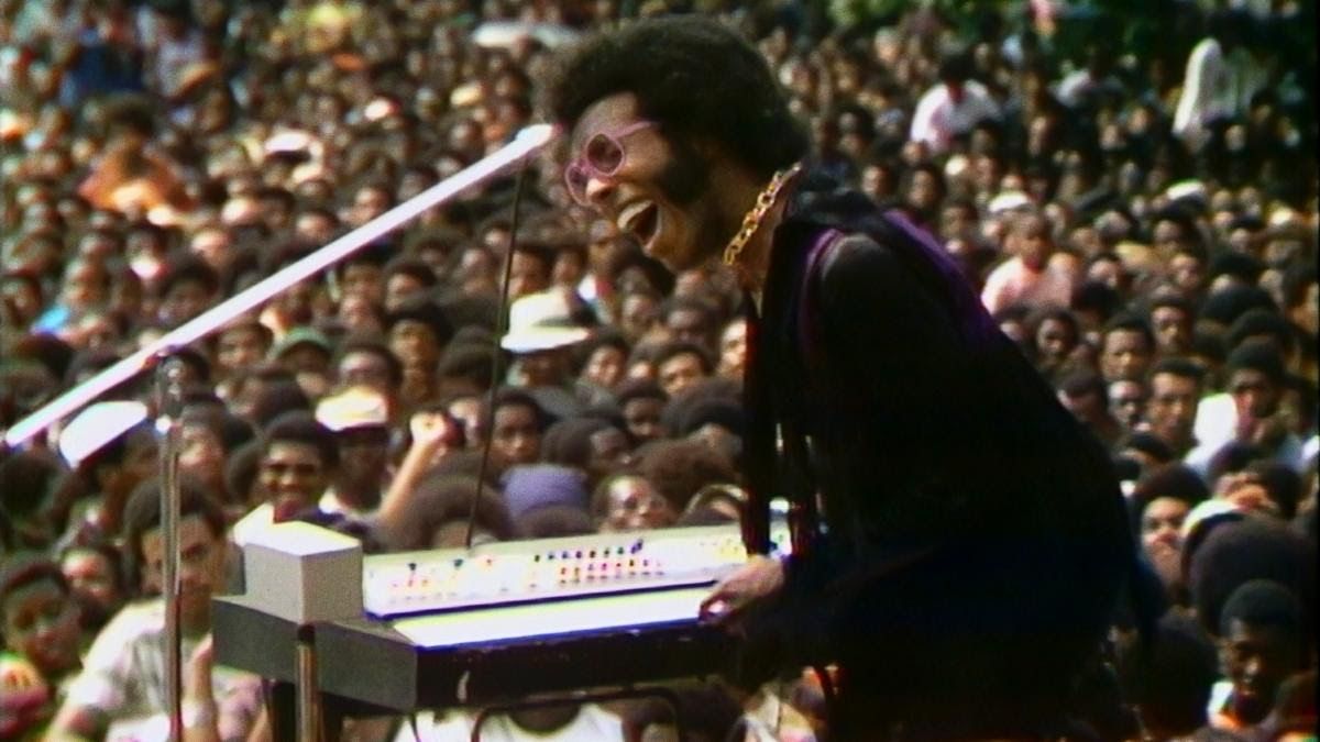 Performer Sly Stone at a music instrument in front of a large, all-Black crowd, enthusiastically performs mid-song