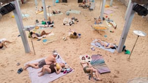 About 20 people dressed for the beach lie on a sandy, brightly lit beachscape created inside a large industrial building.