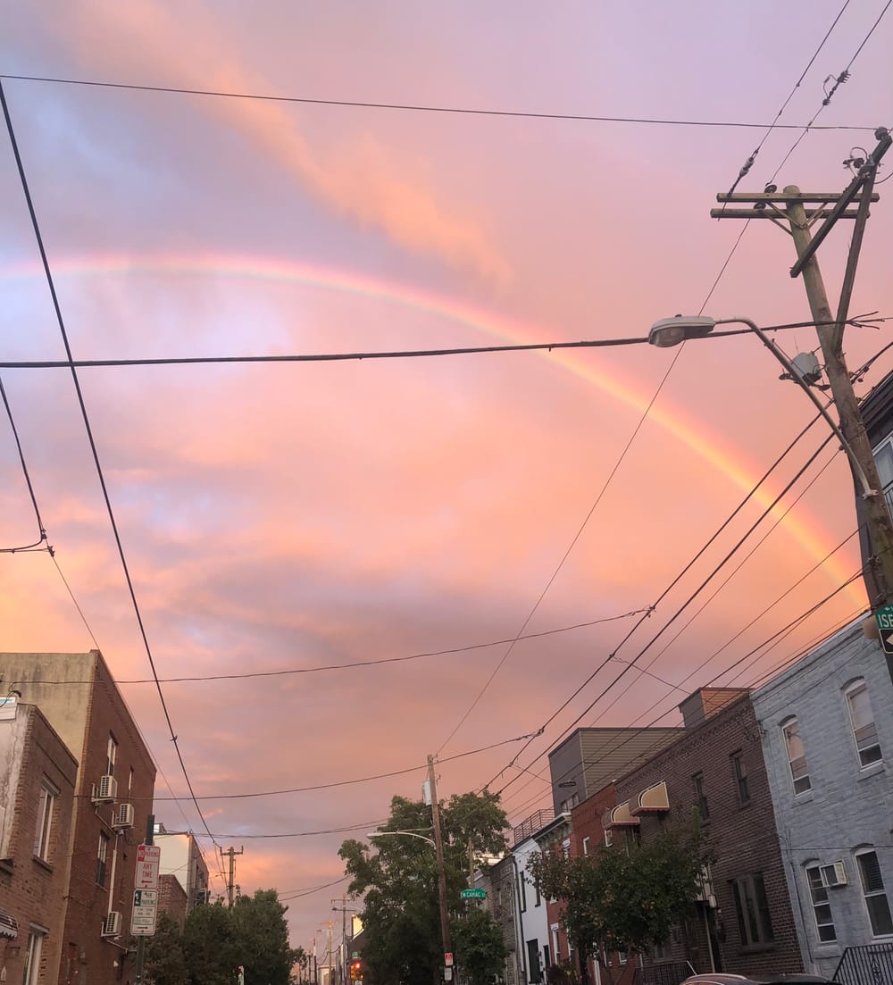 A huge, bright rainbow against a luminous peachy sunset sky over South Philly rowhouses.