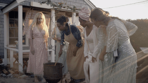 A group of six women outside in gowns outdoors during magic hour, a pot over a fire, Lena stirs with a wooden spoon