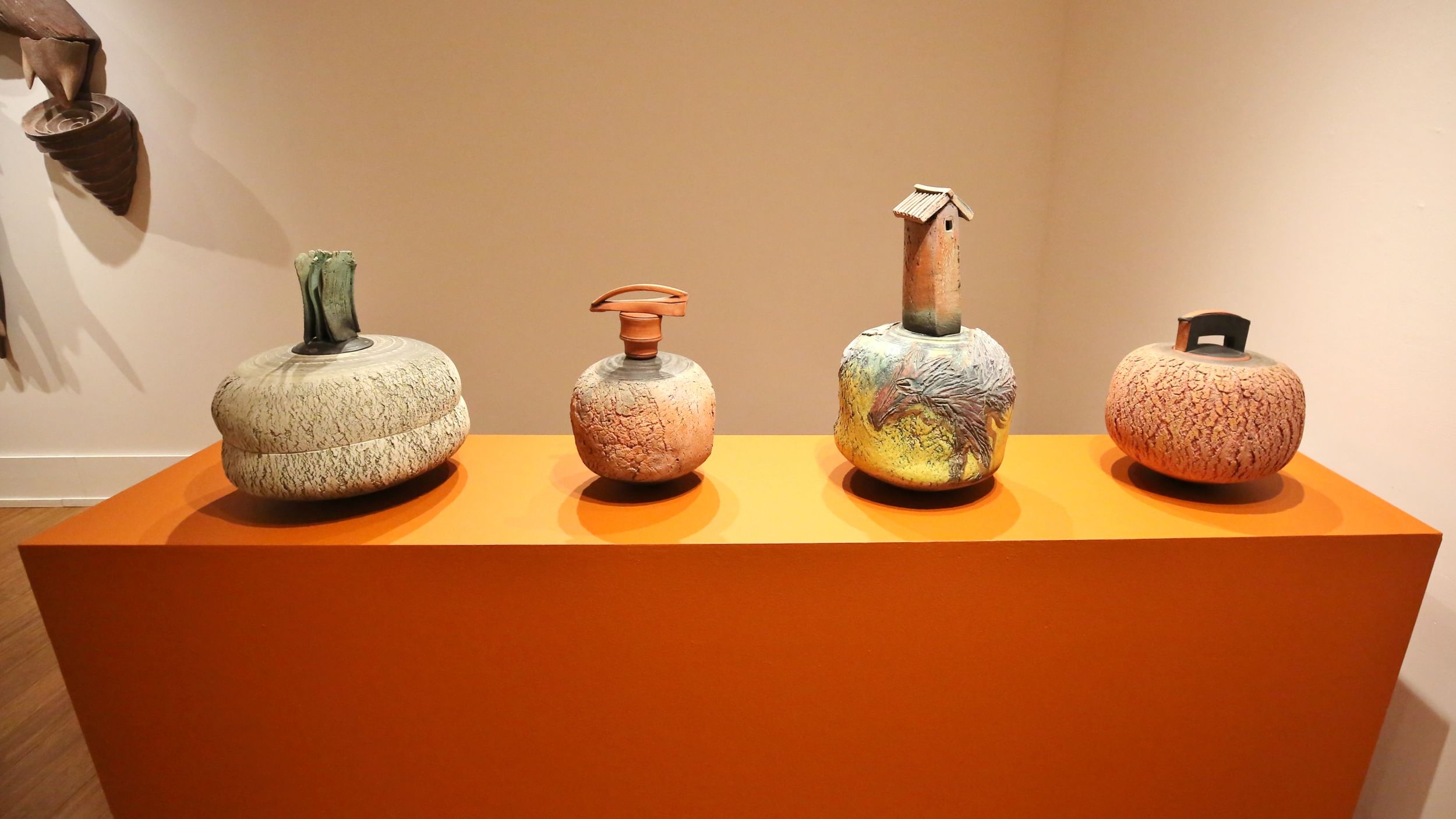 Four rounded ceramic vessels with artful tops and earthy, textured finishes stand in a row on a bright rust-colored base