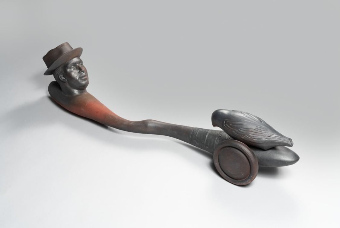 Ceramic sculpture of a man’s head at one end of a long, narrow curving shape with wheels and a bird at the other end.