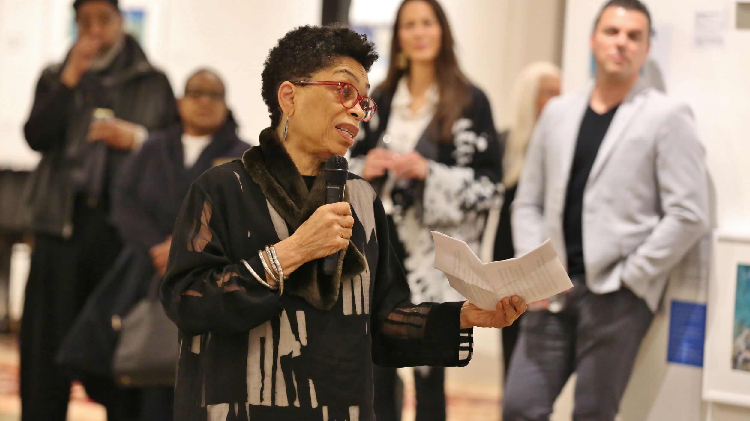 Carpenter, a Black woman with short hair, wearing a textured black & white shirt, speaks into a mic to a crowd in the gallery