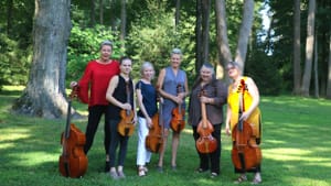Six players each holding a viol pose in a green wooded area with soft sunshine pouring in pockets around them