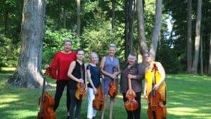 A group of six pose outdoors in a grassy area with tall trees, holding their viols of varying sizes