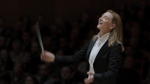 Blanchett as Lydia Tár. She holds a conductor's baton, wears a black jacket w/ white blouse, a crowd out of focus behind her