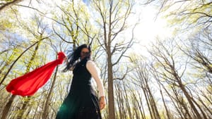 A dancer looks down holding red fabric, camera up at trees with yellow leaves under bright sunlight, other dancers behind her
