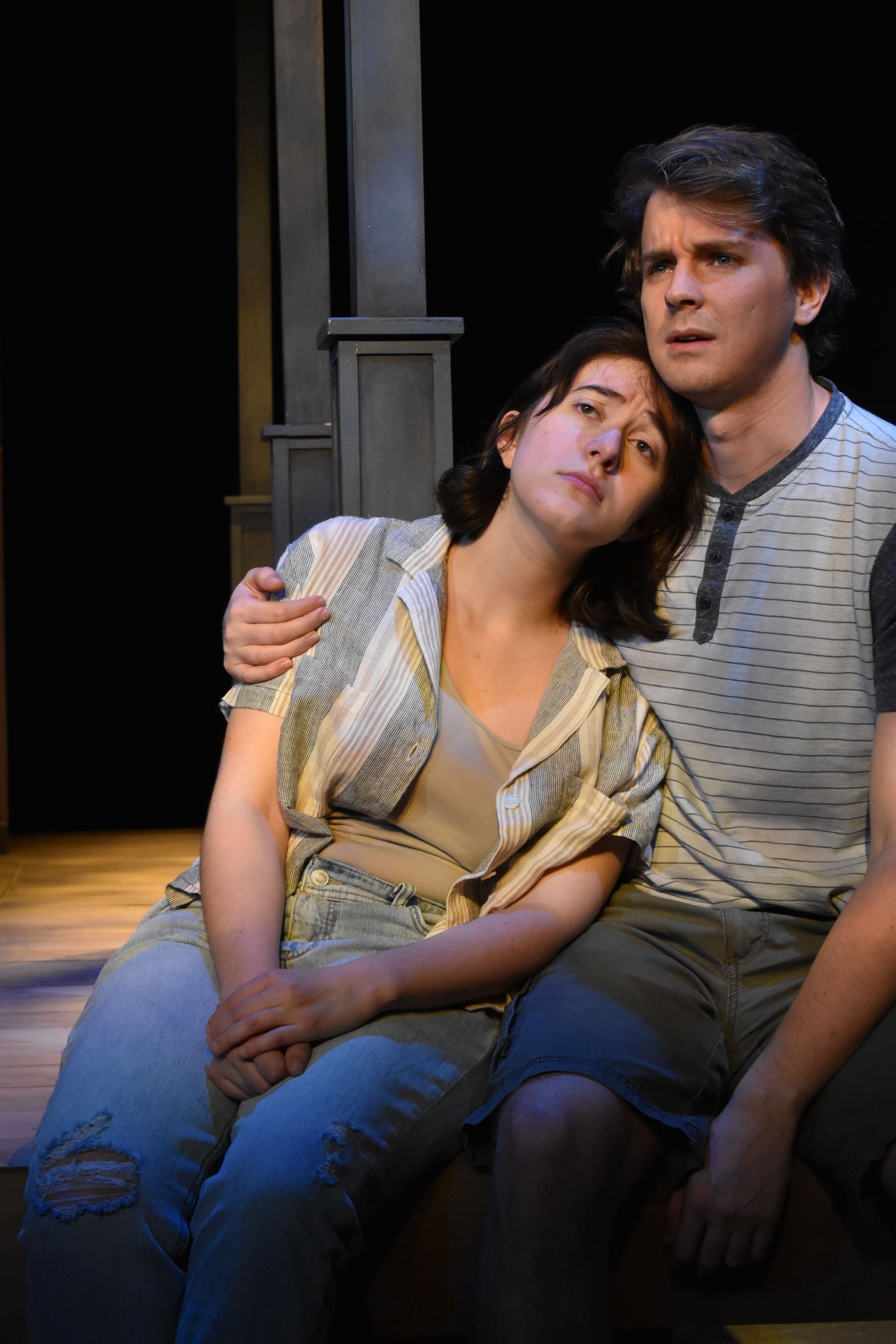 On the edge of the stage, Zion leans sadly against Elmore, who puts an arm around her shoulder. They wear casual clothes.