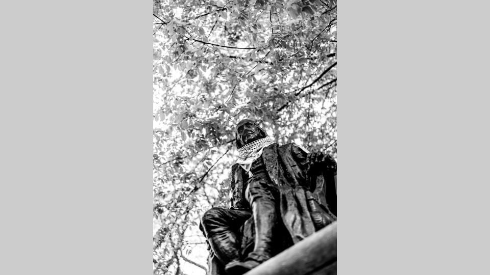 Seen at a sharp angle from below, full-body statue of a seated Ben Franklin has a kefiyyeh on its neck, the tree canopy above