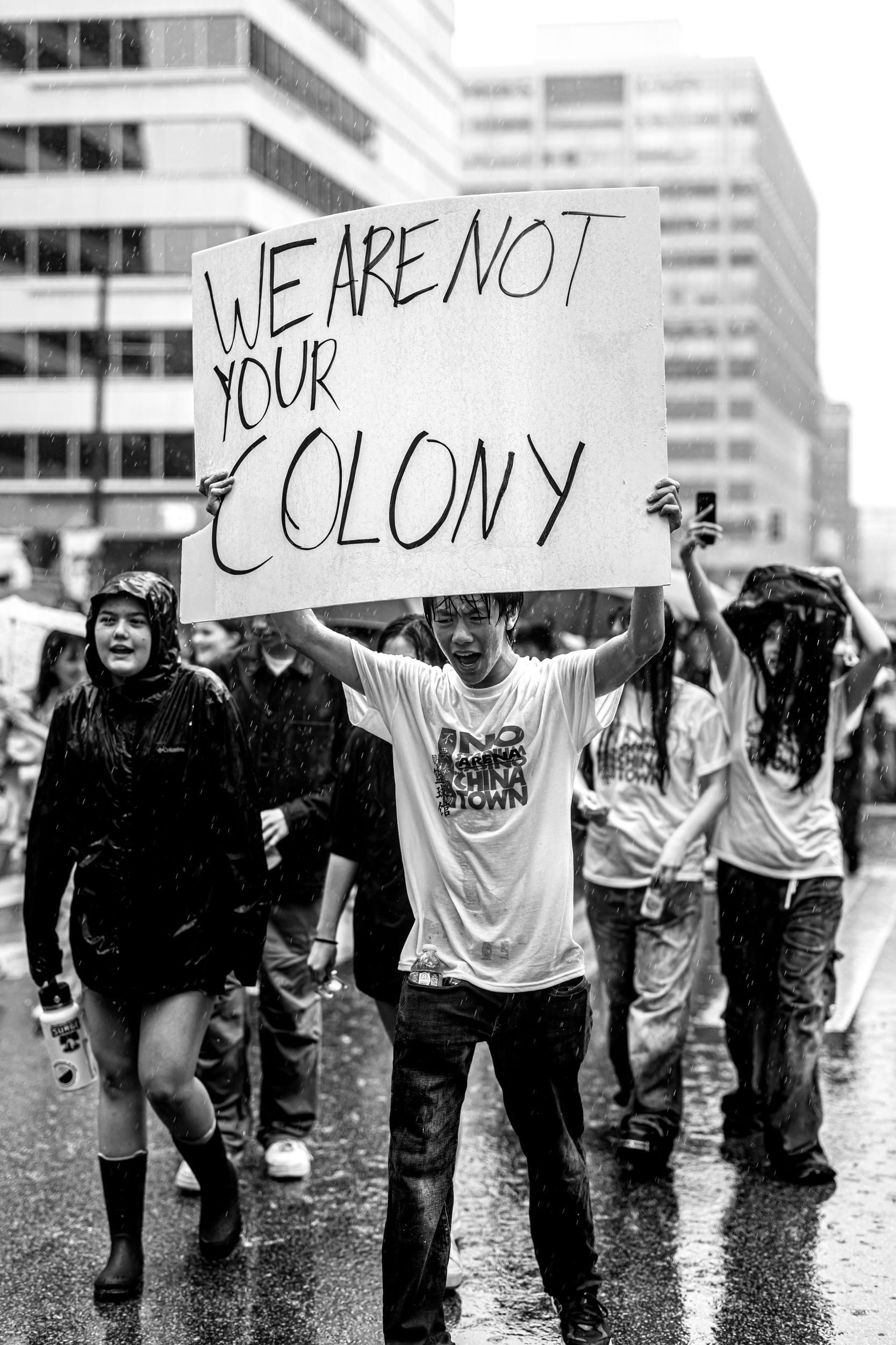 Black & white photo of the head of a protest march in the pouring rain, a young Asian man holding the titular sign, yelling
