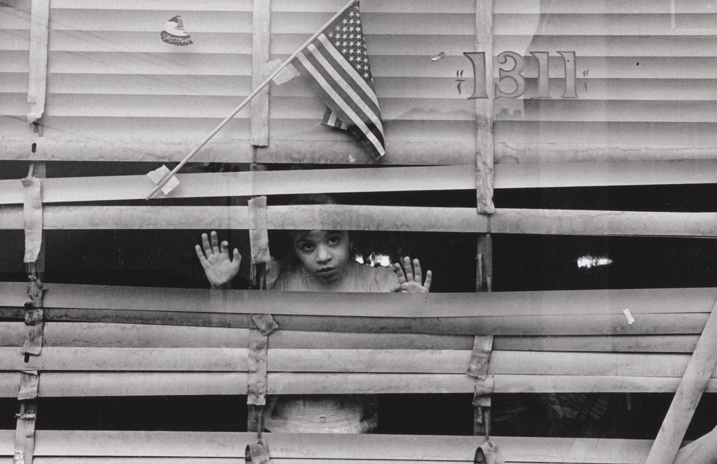Black & white photo of a young child looking out a window between mildewing, broken blinds, a small US flag taped above.