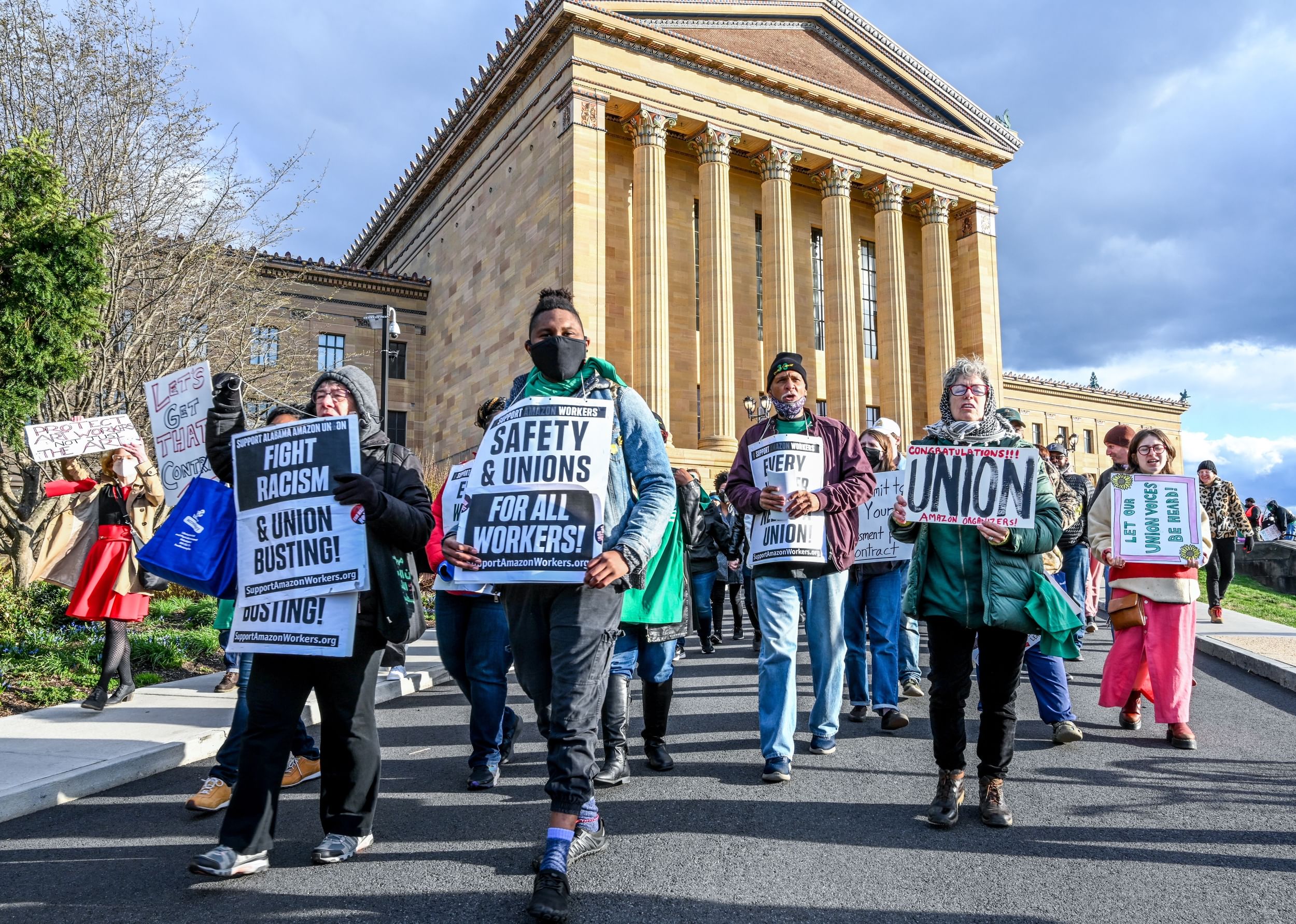 A few dozen people holding pro-labor signs march together on a sunny day below the huge columns of the PMA’s north façade.