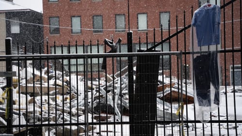 Colored photo of a fenced demolition site in the snow with a brick building behind; a prison uniform hangs at right.