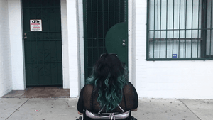 A photo taken from behind of a woman with dark skin and turquoise-tipped hair facing a door too narrow for her wheelchair