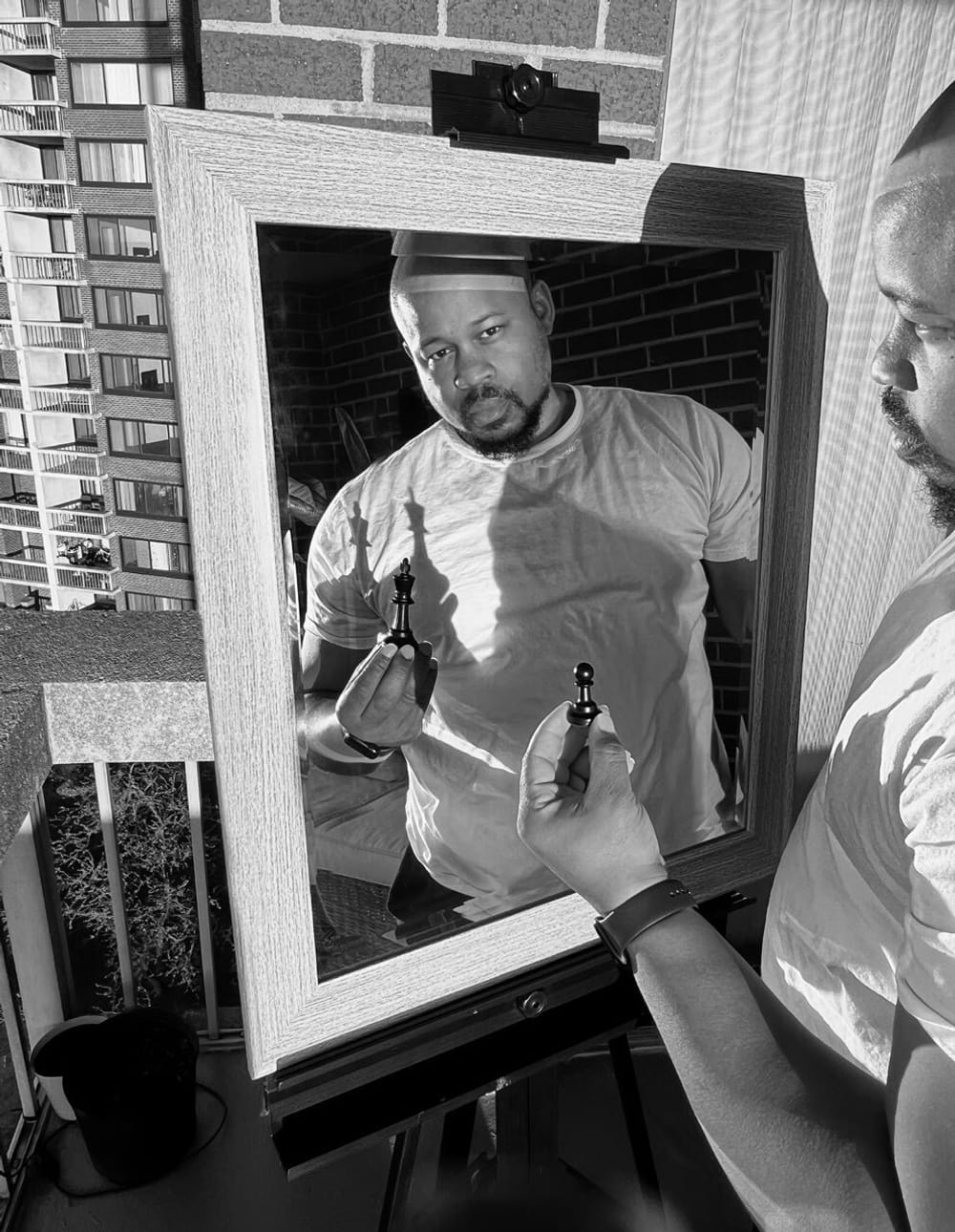Crisp, richly saturated black & white photo of Ray looking in a mirror, holding a chess piece that casts dramatic shadows