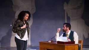 Scene from the play. Banks stands warily opposite Close, seated at a desk in vest & tie. She wears a camo jacket & a backpack