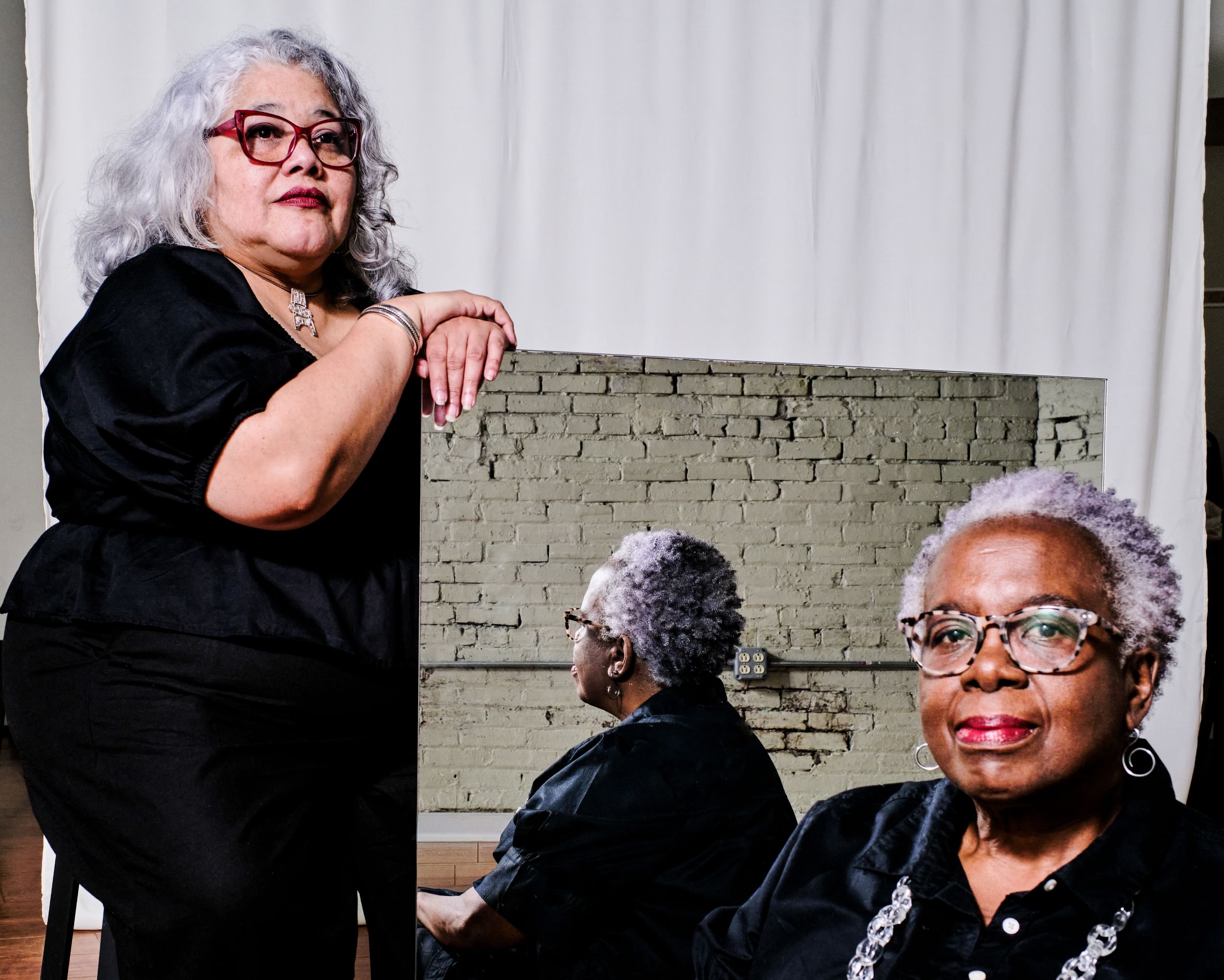 Two women in black, with gray hair, pose for portrait. One looks at camera, her image reflected in a mirror held by the other