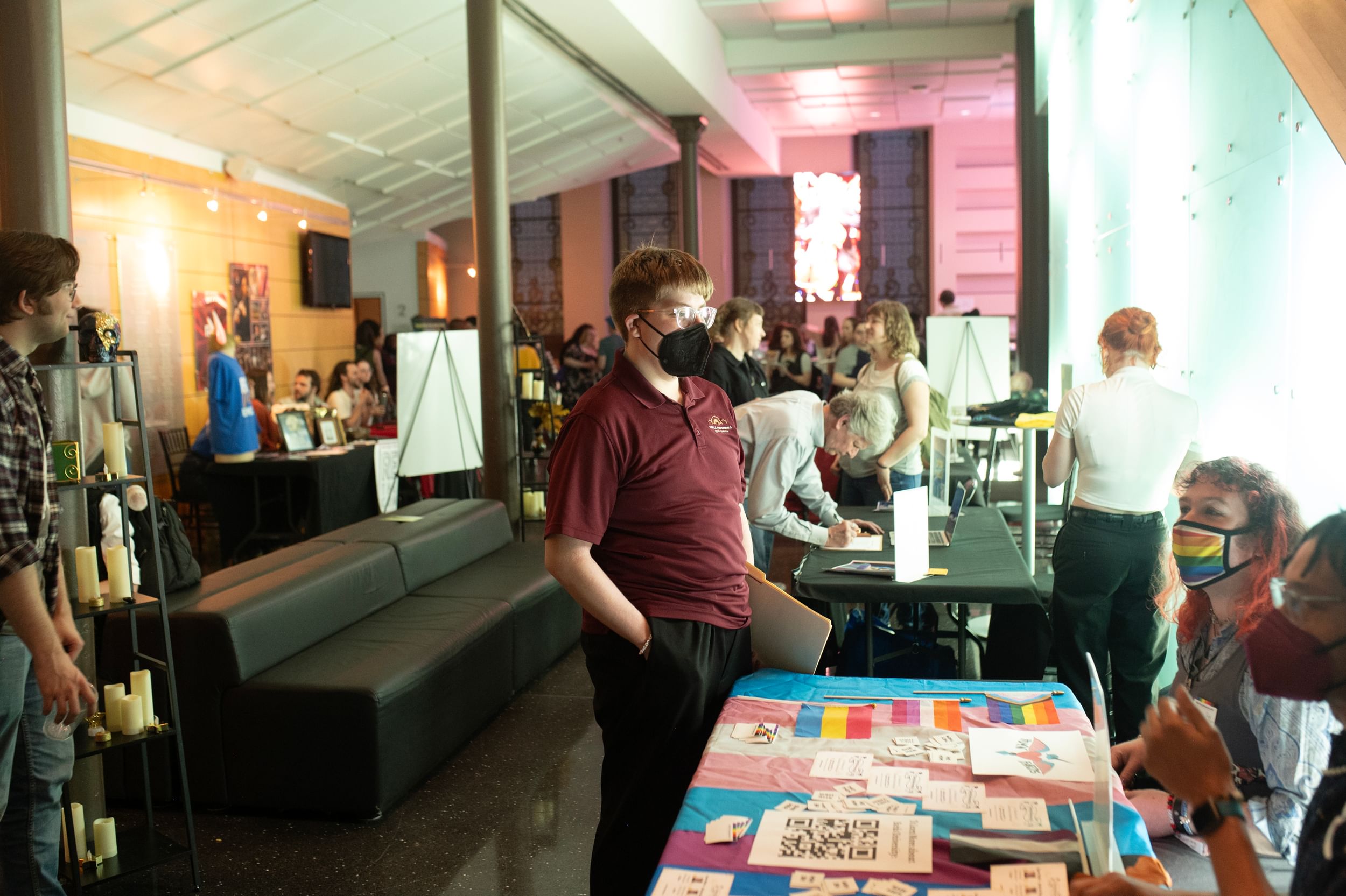In a small lobby, people, some of them masked, interact over decorated tables.