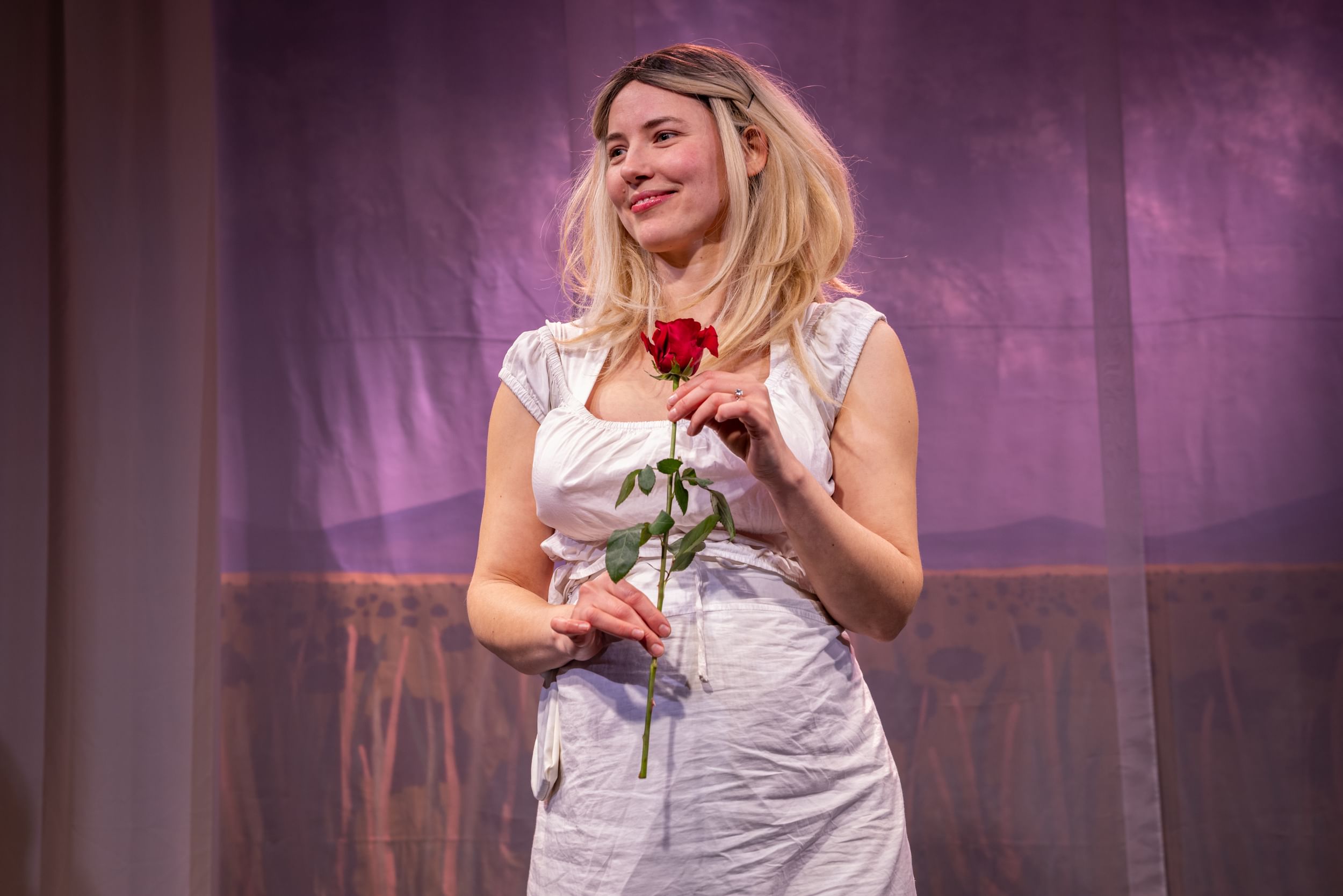 Mitchell, a white woman in a blonde wig, white dress, and fake pregnant belly, smiles while holding a rose.