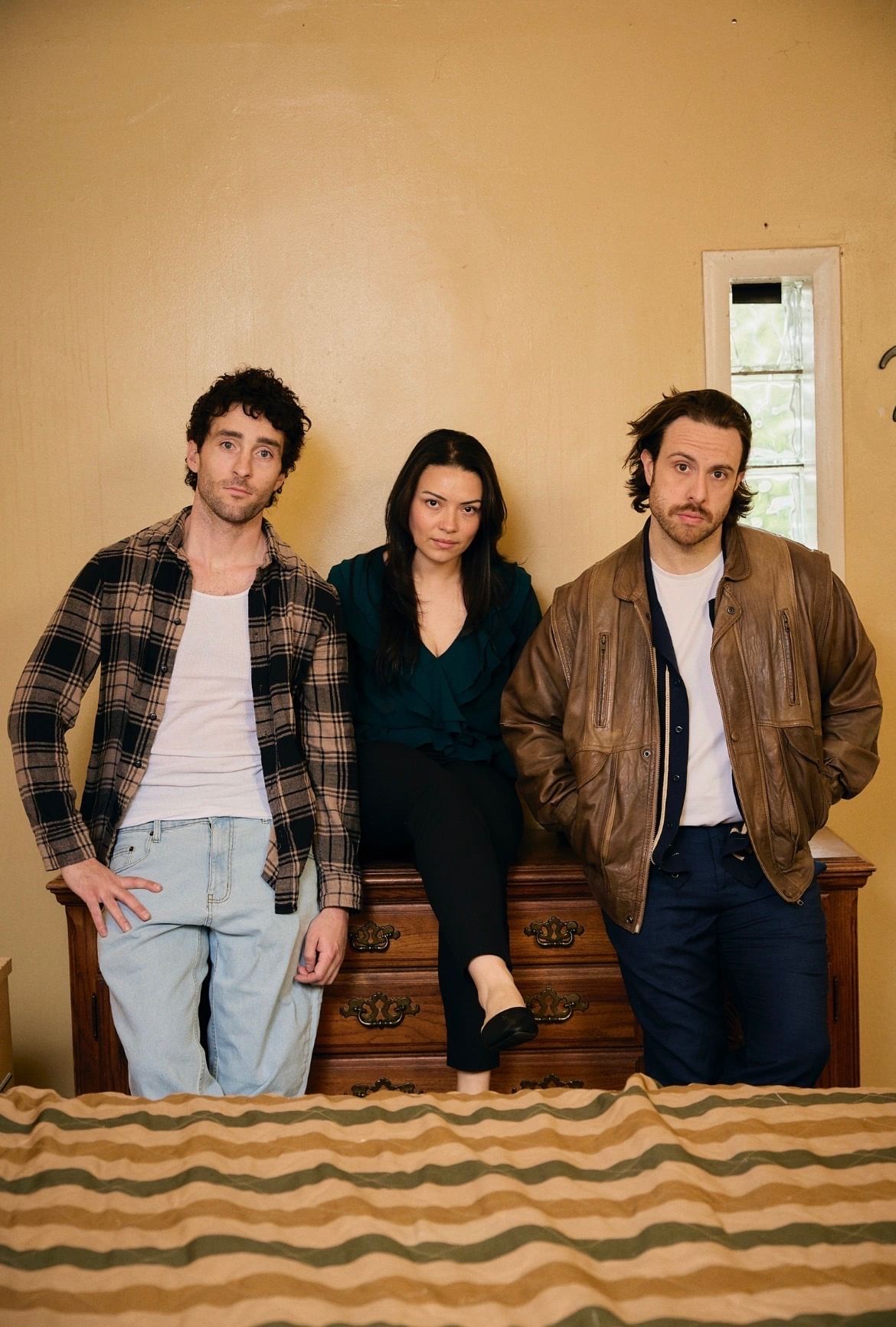Three white actors in their 20s in casual clothes lean or sit on a dresser close together, looking portentously at the viewer