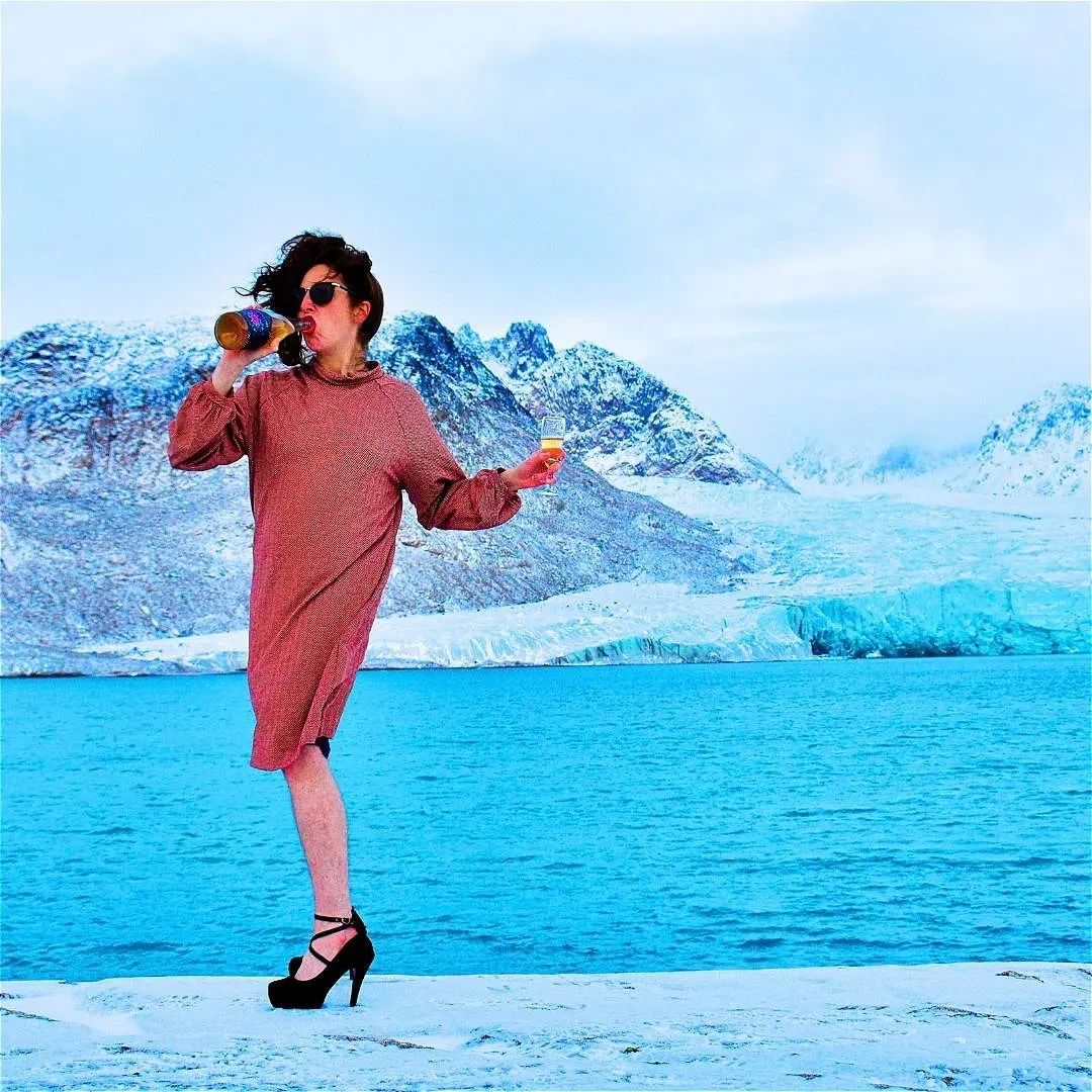 A person drinks from a bottle, holds champagne glass in other hand, wearing a dress and heels in an arctic area