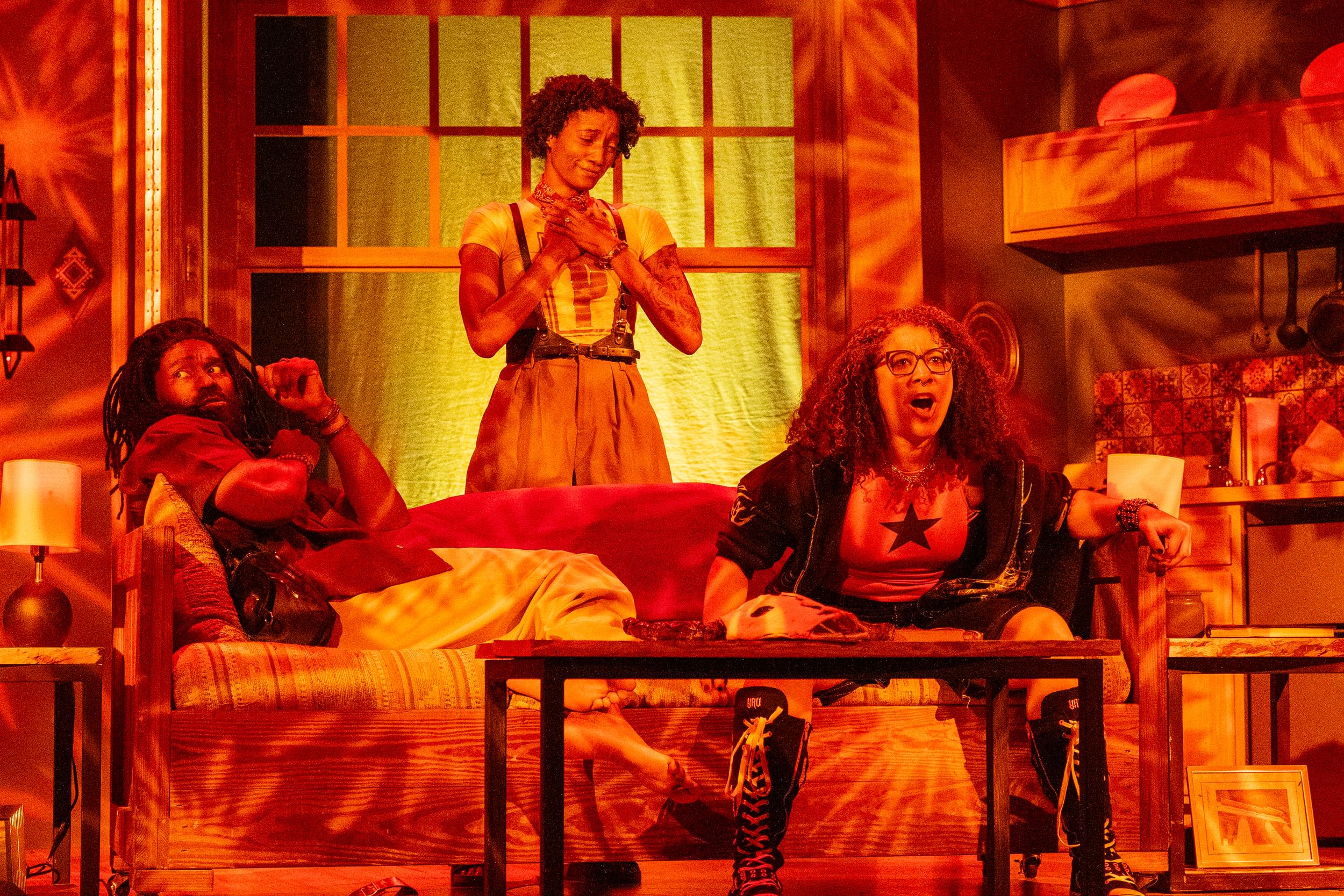 In orange lights, DeShields looks scared lounging on a couch by Cox, looking shocked. Hall stands behind, hands on her chest