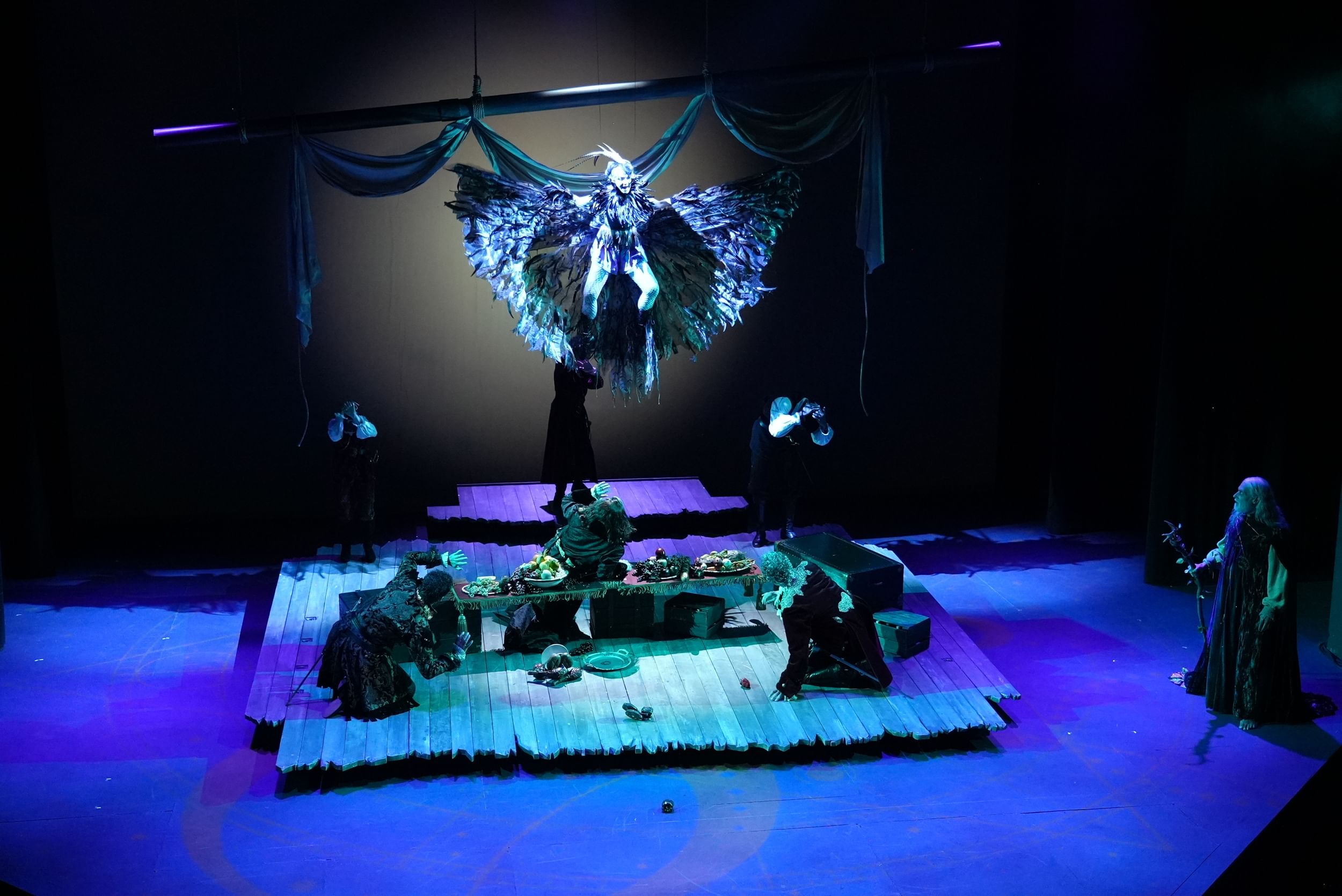 Gliko, in a fantastical feathered costume, descends over five feasting, terrified sailors on a dramatic blue-lit stage.